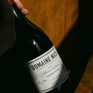 Mano sujetando del cuello una botella de vino Domaine Nico Grand Mère Pinot Noir 2019 sobre un fondo de madera.