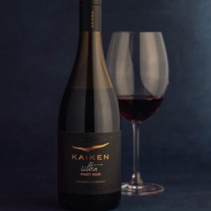 Botella de vino tinto Kaiken Ultra Pinot Noir junto a una copa servida sobre un fondo azul oscuro.