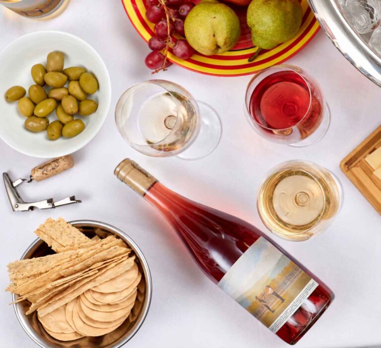 Botella de Amiguito Criolla de Bodega Aleanna sobre una mesa con copas de vino, aceitunas, frutas y galletas, vista desde arriba.